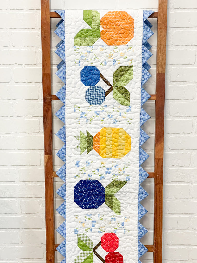 Ambrosia Table Runner. Blue triangle border with variety of fruits quilted onto white background 