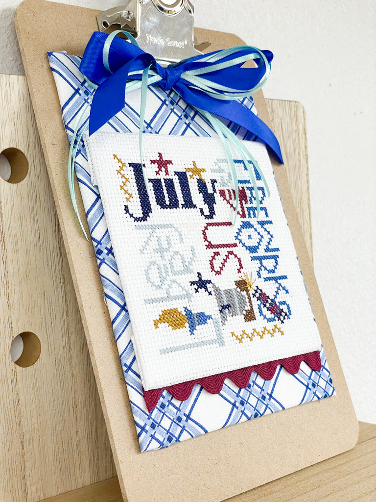 Pine Mountain Design July cross stitch on a clipboard with two blue bows