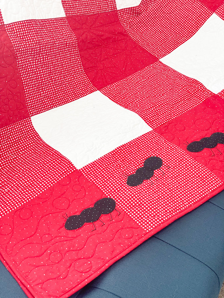 Red and white gingham quilt with three quilted black ants on the bottom corner 